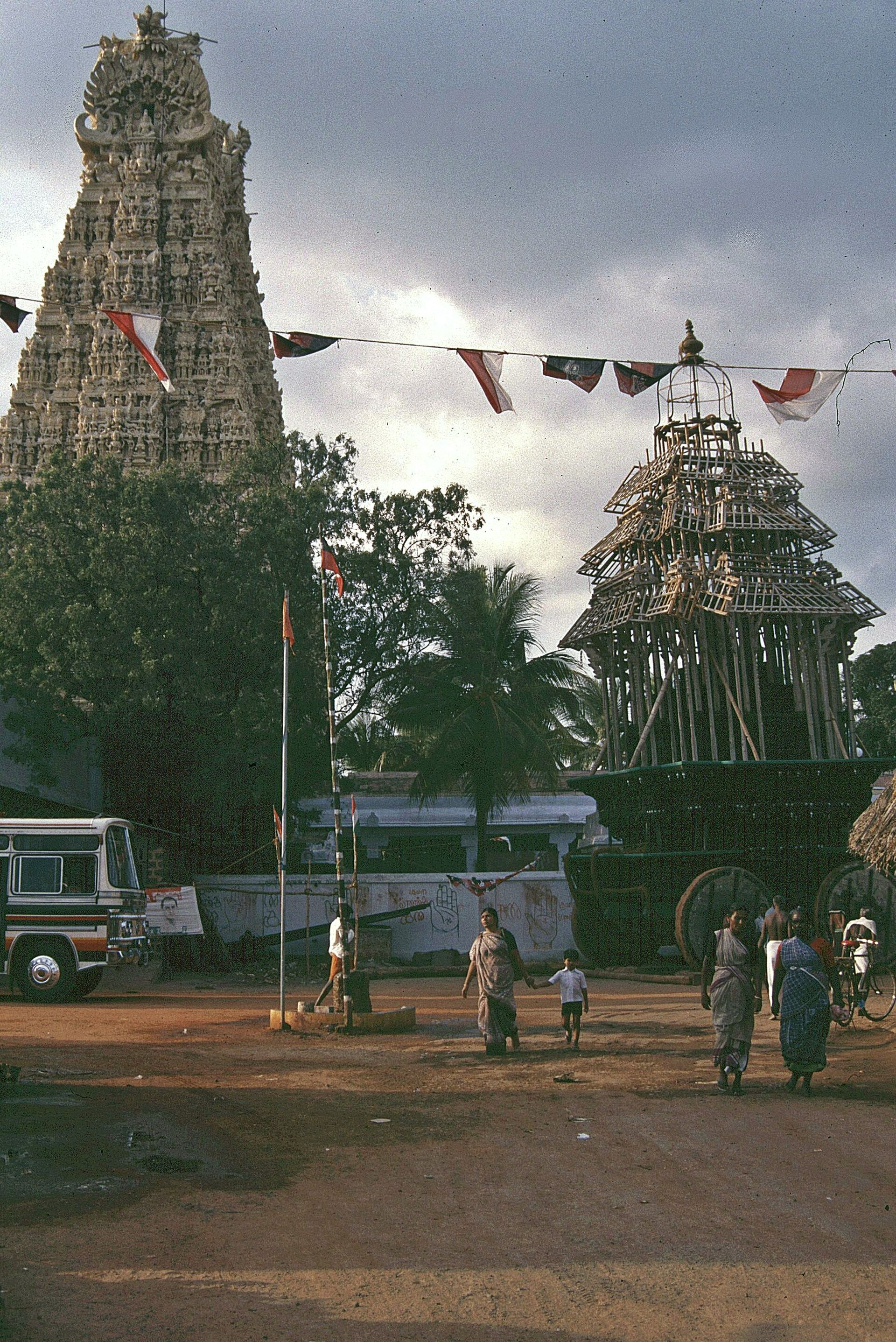 Color photograph from 1989 of the Suchindram temple gopuram beside a tall wooden temple car, with people and a bus in the street.